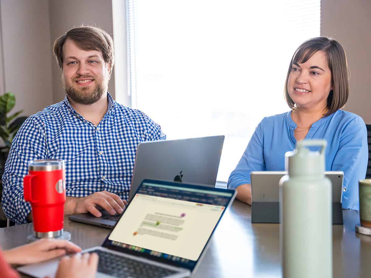 Two employees in a meeting smiling at a third employee off-camera whose laptop screen shows a collaboration document.