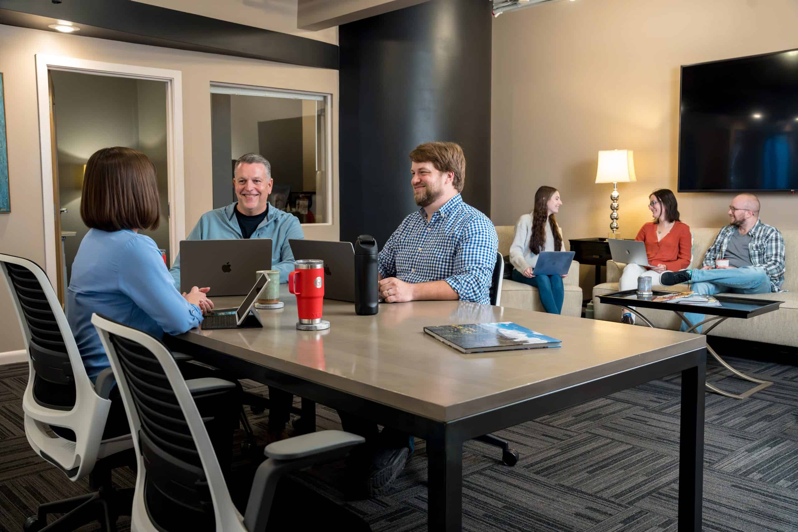 A group of employees collaborating at a conference table and another group in the background collaborating on a couch and chair